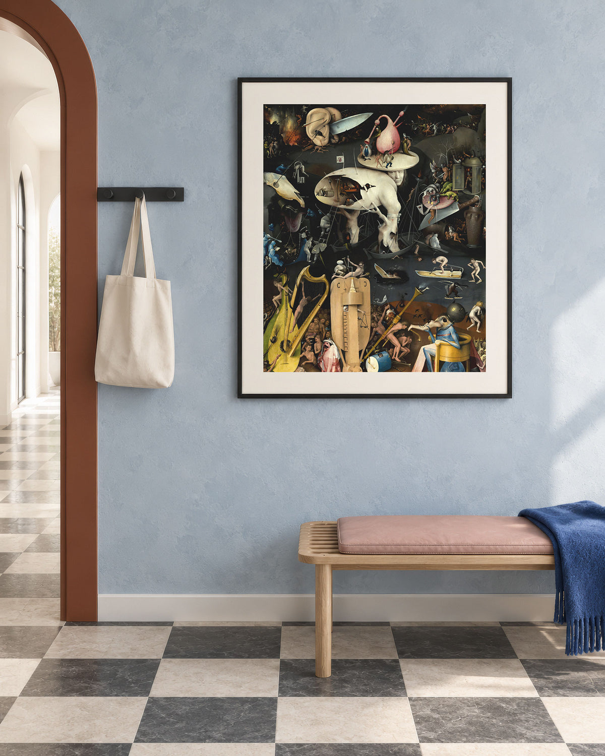 A framed surreal art print hangs on a light blue wall in a styled entryway with a bench, blue throw blanket, and a cream tote bag hanging on a hook.