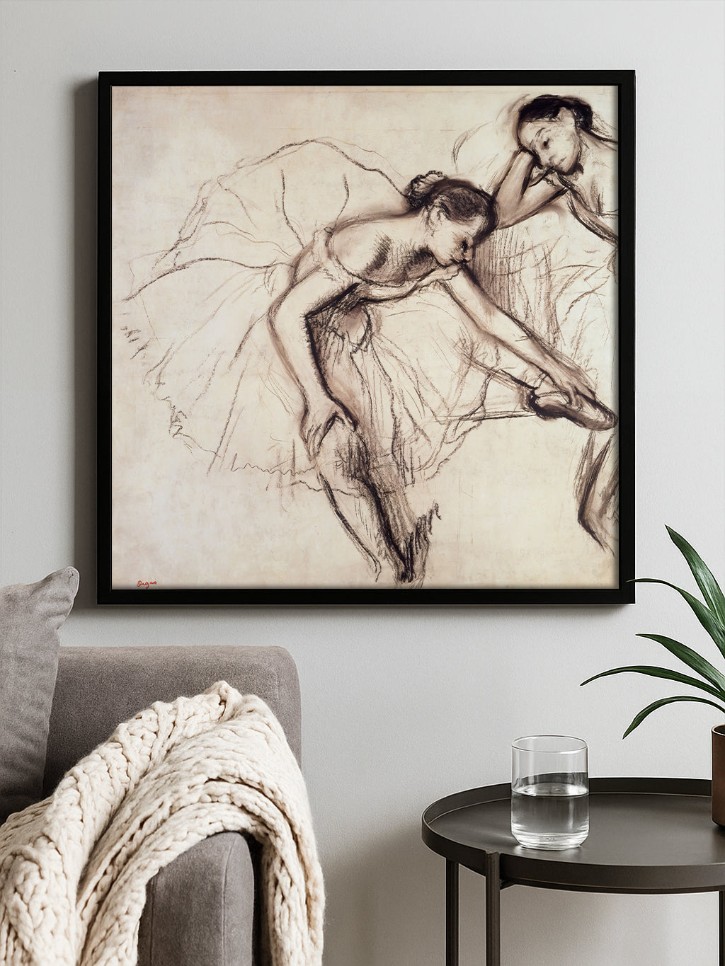 A framed art print featuring a sepia-toned sketch of two ballerinas, displayed on a light gray wall above a gray sofa with a chunky knit throw blanket and a small side table with a glass of water and a plant.