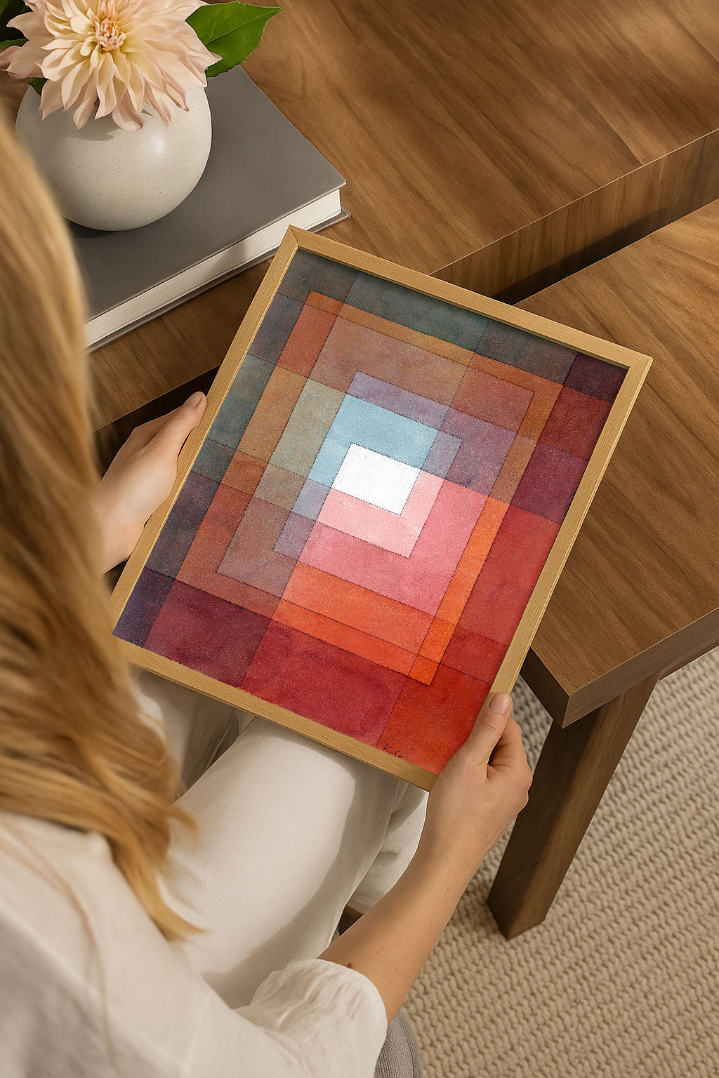 A person holds a square framed art print featuring concentric geometric squares in shades of red, orange, pink, and blue. The scene is set in a warm, modern room with a wooden table and a vase of flowers in the background.