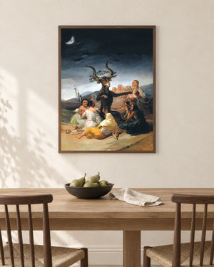 A framed classical painting hanging on a beige wall above a wooden dining table with chairs and a bowl of pears. The painting depicts a dark, dramatic scene with several figures and a horned figure under a crescent moon.