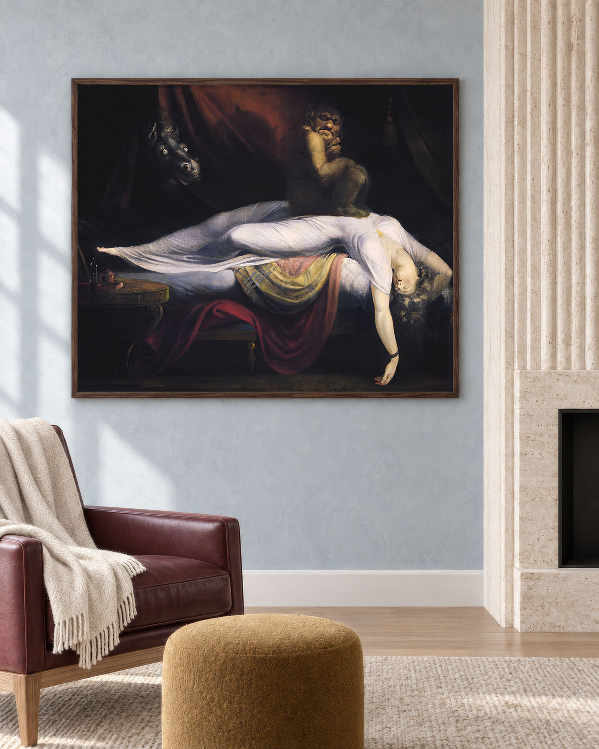 A framed classical painting depicting a dramatic scene with figures and flowing red and white drapery, displayed on a light blue-gray wall in a styled living room with a leather armchair, ottoman, and fireplace.